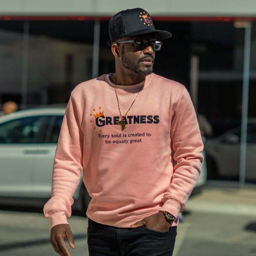 Black man wearing a pink sweatshirt with 'GREATNESS' text, black cap, and sunglasses outdoors.