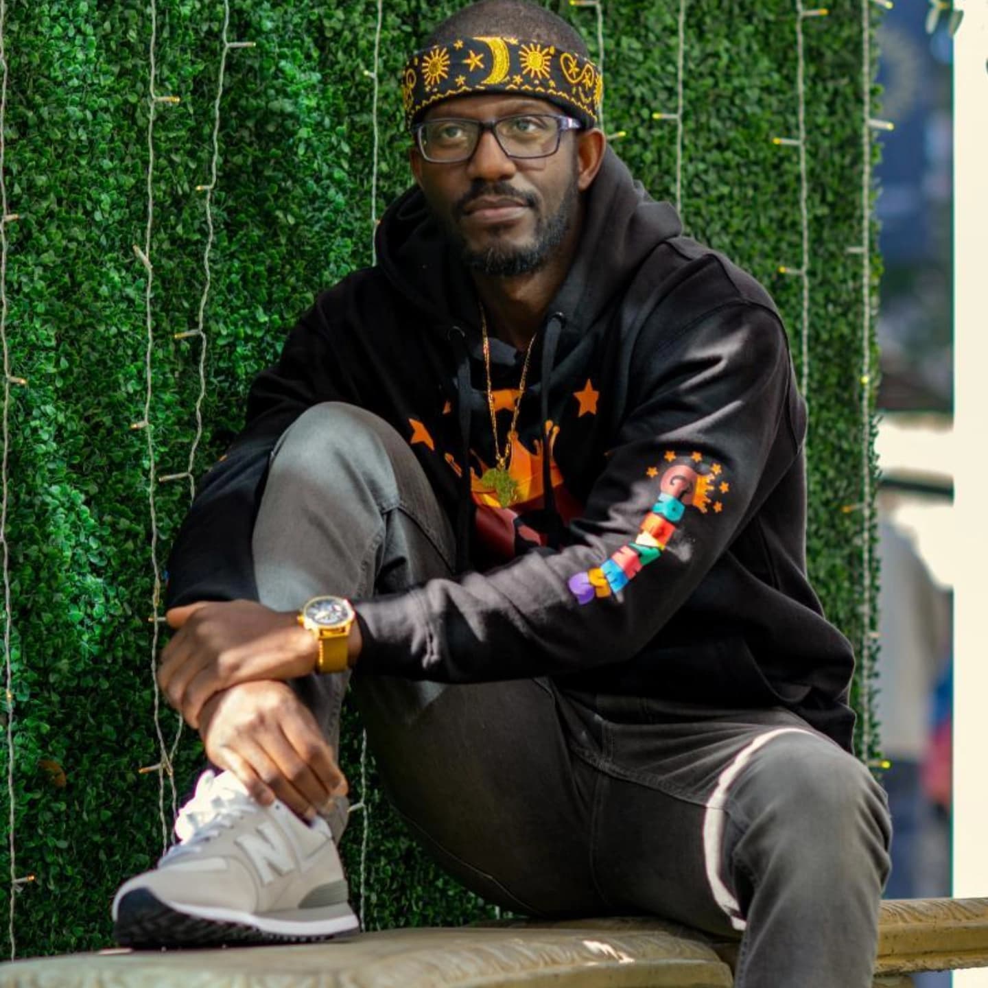 Man wearing a celestial headband and black hoodie sits against a green hedge wall.